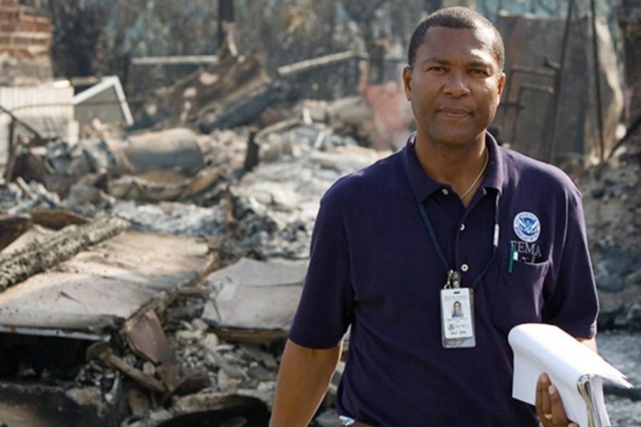 FEMA official standing in a disaster-affected area, holding documents while assessing damage from a wildfire or other destructive event.