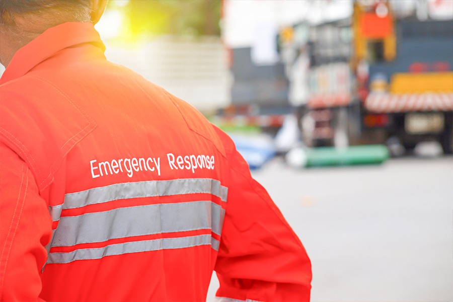 Emergency response worker in a high-visibility jacket at an incident scene, with emergency vehicles and equipment in the background.