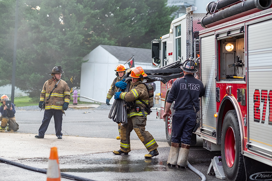 A group of firefighters in full turnout gear work near a red and white fire engine. One firefighter carries a bundled hose on their shoulder while another person wearing a dark shirt labeled "DFRS" stands at the back of the truck.