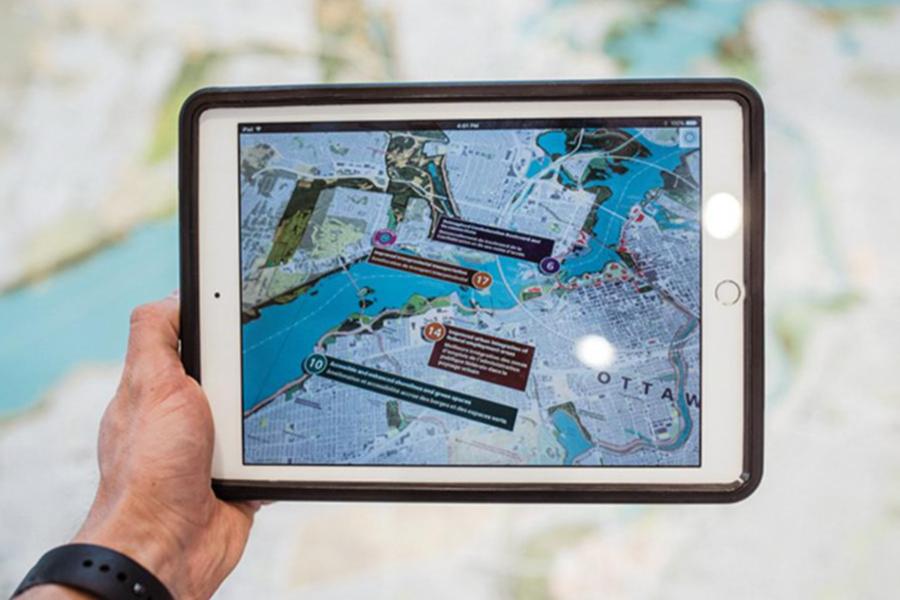A person holds a white tablet displaying an interactive, augmented-reality digital map of Ottawa over a larger printed paper map. The digital screen shows the city's river and streets overlayed with numbered, dark text boxes providing localized information.