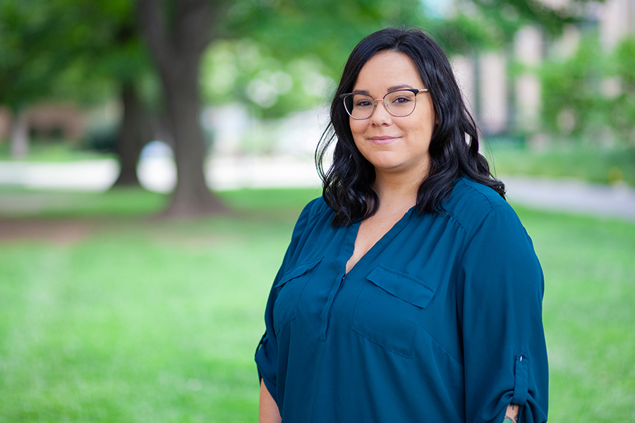 A smiling woman with shoulder-length dark hair and glasses poses outdoors in front of a blurred green park background, wearing a teal blouse.