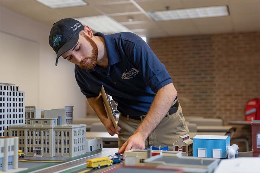 A student reviews a tabletop disaster response simulation. He is positioning model school buses and cars in a miniature city layout featuring school and industrial buildings.