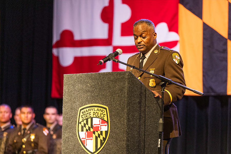 A Maryland State Police officer speaks at a podium with the department's emblem. Flags are in the background; the atmosphere is formal and official.