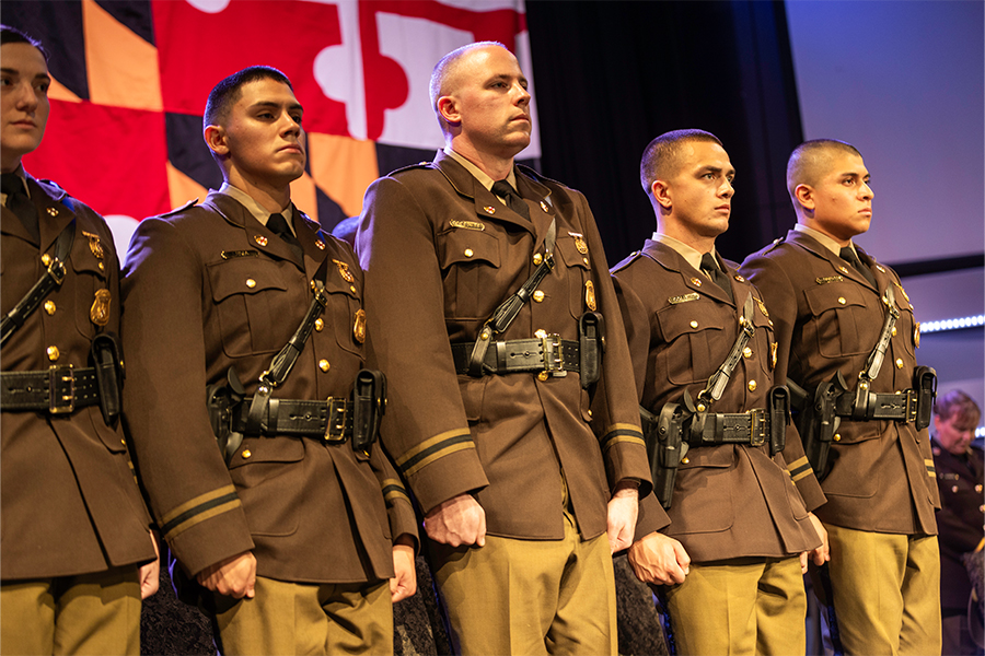 A row of five solemn state troopers in brown uniforms stand at attention on stage, against a backdrop resembling the Maryland flag.