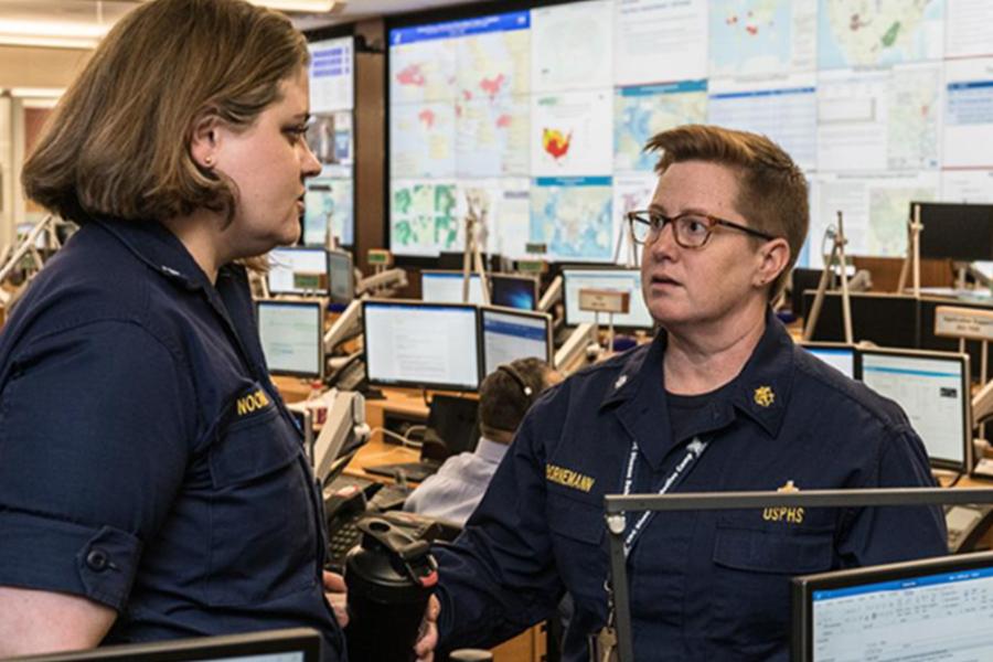 Emergency operations personnel discussing response activities in a command center, with computer workstations and monitoring screens in the background.