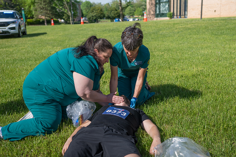 Two medical responders in teal scrubs kneel on a grassy field to assist a person lying flat on their back, who is wearing a black shirt with a race bib numbered 8373.