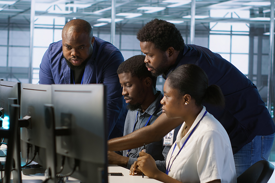 A group of four professionals look intently at computer monitors in a modern office or server room setting.