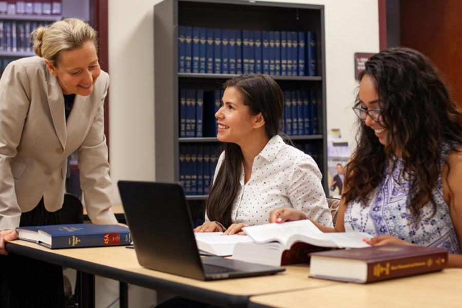 A law professor or librarian stands next to two smiling female students who are studying with thick legal books and a laptop in a law library setting.