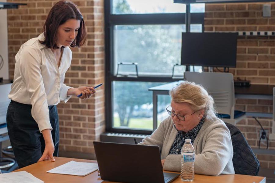 A younger woman standing in a white blouse looks over the shoulder of an older woman with glasses who is sitting at a wooden desk and working on a laptop.