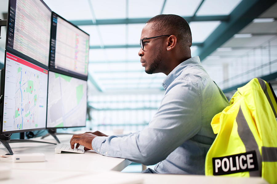 Police operations center employee monitoring multiple computer screens displaying maps and data, with a high-visibility police jacket on a nearby chair.