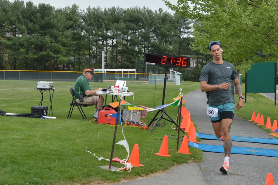 A focused male student-athlete with bib number 56 runs past a digital race timer and a timing official on a campus path near athletic fields.