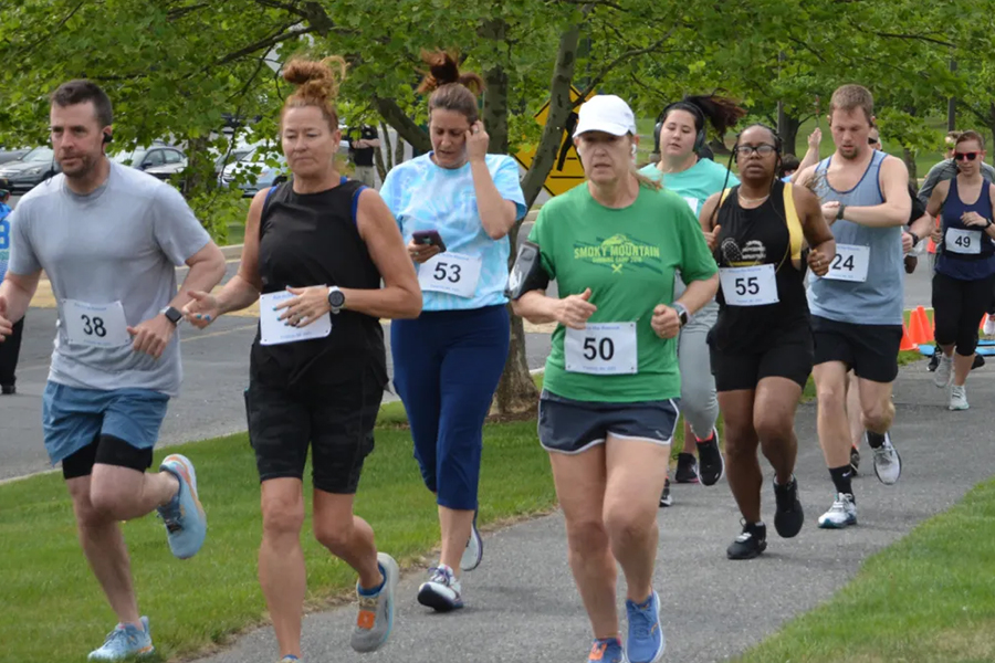 Group of adult runners participating in a community road race or 5K run on a tree-lined sidewalk in a park-like suburban setting. Several men and women wearing athletic clothing, running shoes, and numbered race bibs jog closely together during the event.
