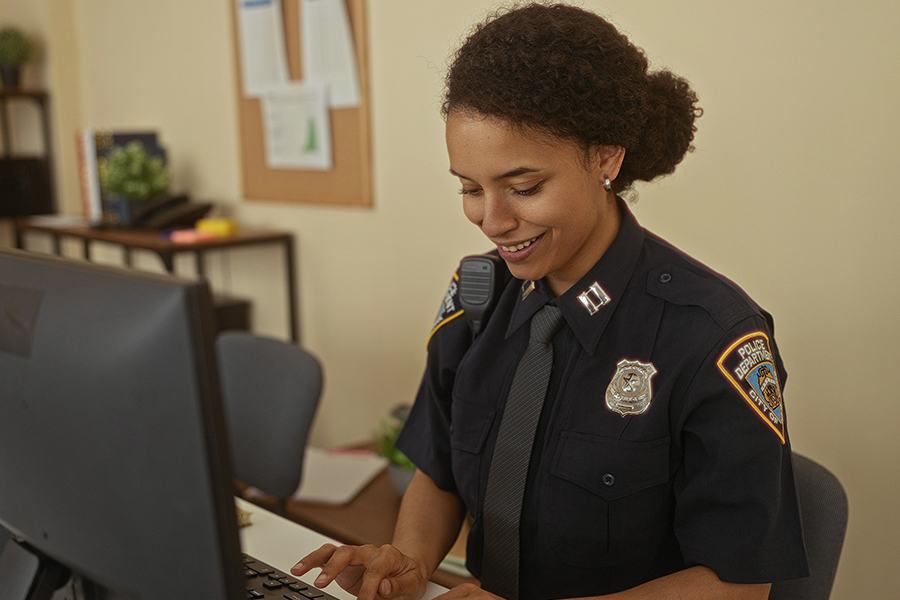 A smiling police officer in uniform types on a computer keyboard in an office setting. The backdrop includes a bulletin board and a shelf with books, conveying a professional and focused atmosphere.