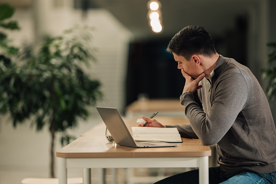 A man sits at a wooden table engrossed in his work, pen in hand, with a laptop and notebook. The space is calm, featuring soft lighting and greenery.