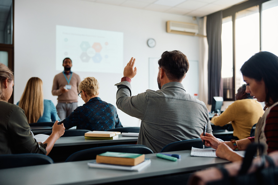 Participants sit in a classroom during a training session while a presenter speaks at the front of the room. One attendee raises a hand to ask a question, while others take notes and listen.