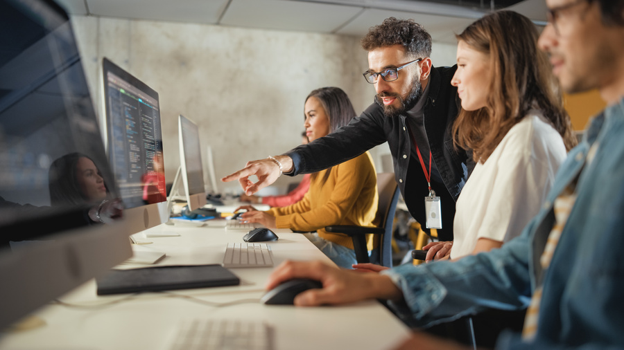A team of professionals works together at computer workstations in a modern office. One man leans over and points to information displayed on a monitor while colleagues watch and interact with their computers.