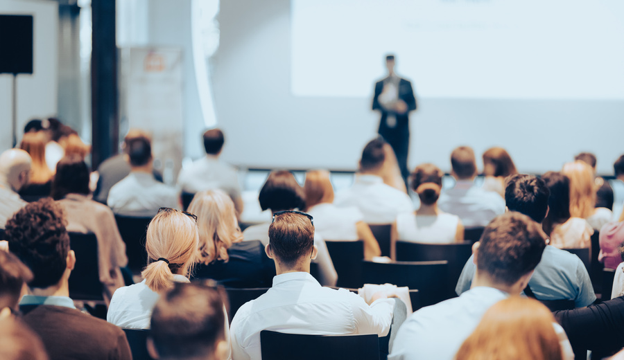 Attendees sit in a conference audience facing a presenter speaking at the front of the room during a professional event or seminar.