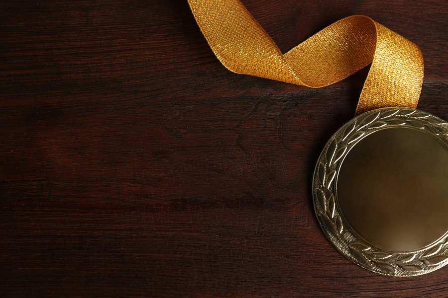 A gold medal with a ribbon rests on a dark wooden surface, viewed from above. The medal is positioned in the lower-right corner, leaving ample empty space across the image.