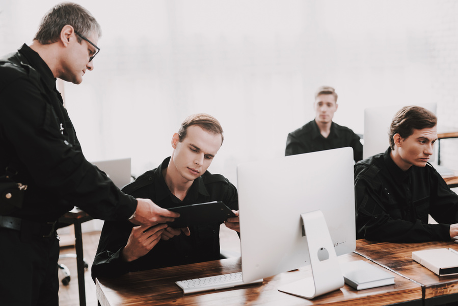 Several security or law enforcement personnel work at computer stations while reviewing information on a tablet in a training or operations environment.