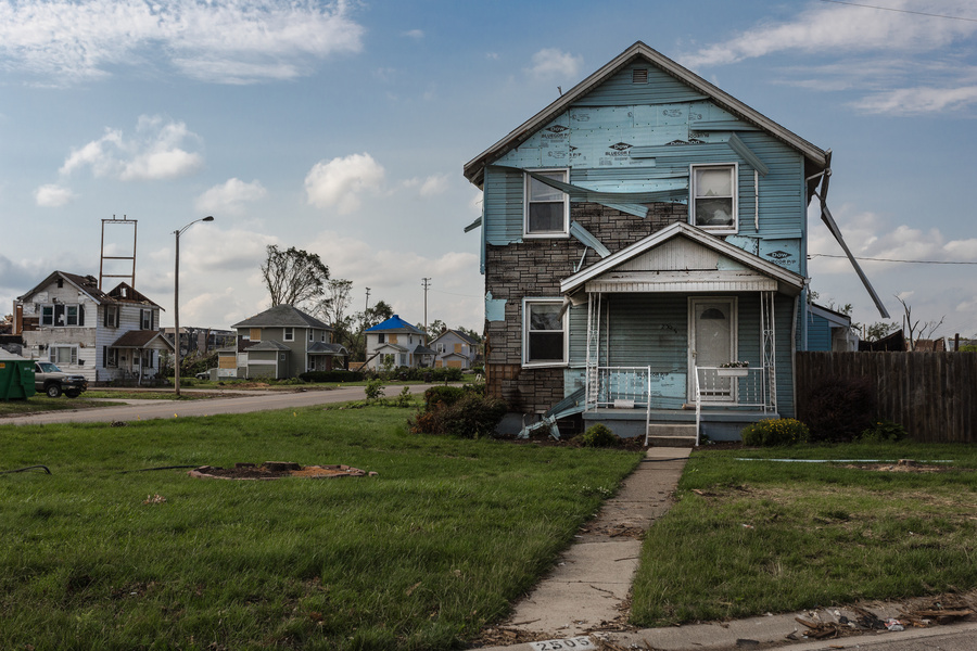 A weathered two-story house with visible structural damage stands in a neighborhood recovering from a disaster. Damaged buildings and debris can be seen in the surrounding area.