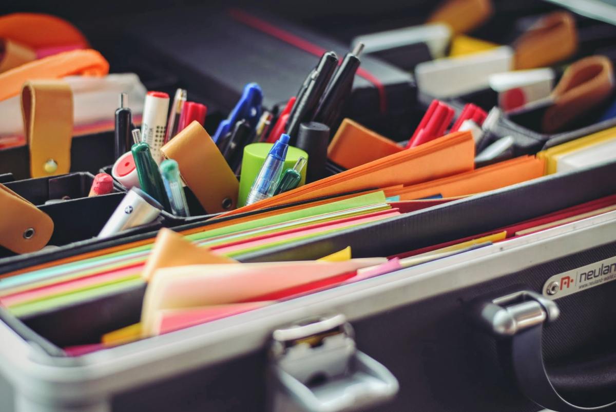 An open, black briefcase style organizer is tightly packed with a variety of colorful office supplies, including pens, markers, scissors, and stacks of multicolored cards.