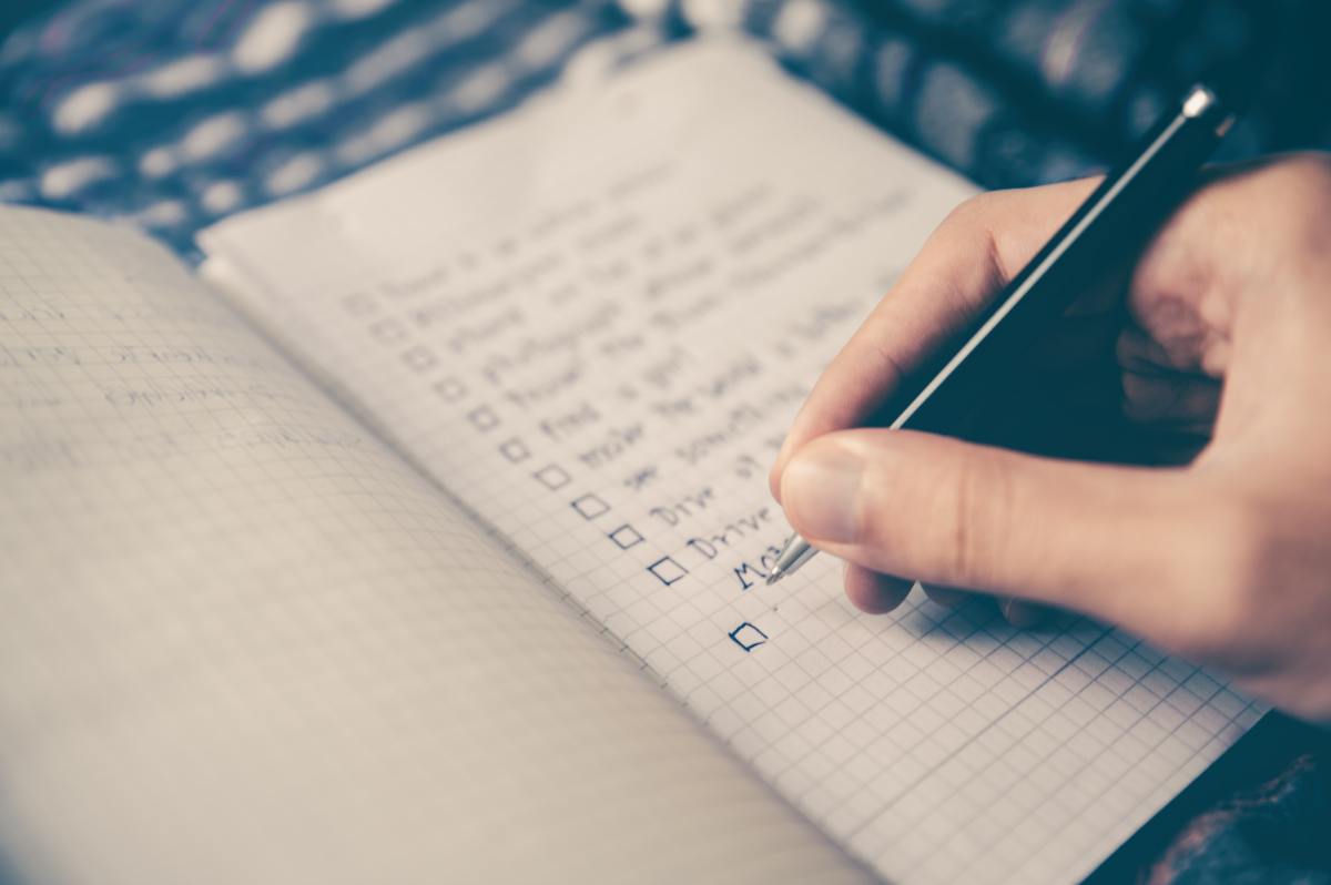 A close-up of a person's hand writing with a black pen on a checklist in a grid-paper notebook.