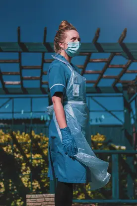 A healthcare worker wearing a blue uniform, face mask, blue gloves, and a clear plastic apron looks off to the side while standing outdoors in front of a wooden pergola structure.