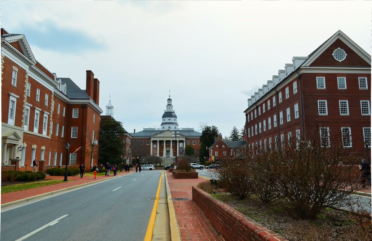 A paved road flanked by large brick academic buildings leads toward the historic Maryland State House with its prominent white dome under a cloudy sky.