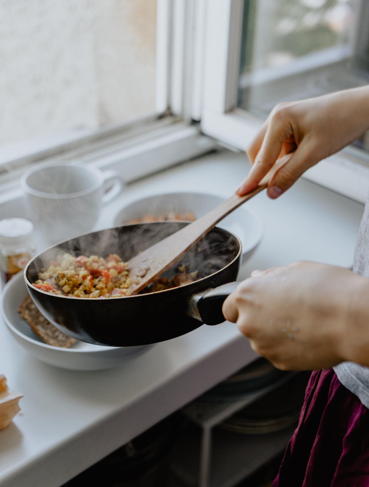 A person uses a wooden spatula to stir steaming food in a black skillet held over a white windowsill.