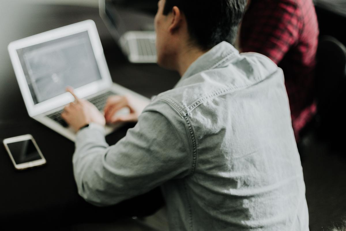 A person wearing a light grey denim shirt sits at a dark desk and points at an open laptop screen with a smartphone resting nearby.