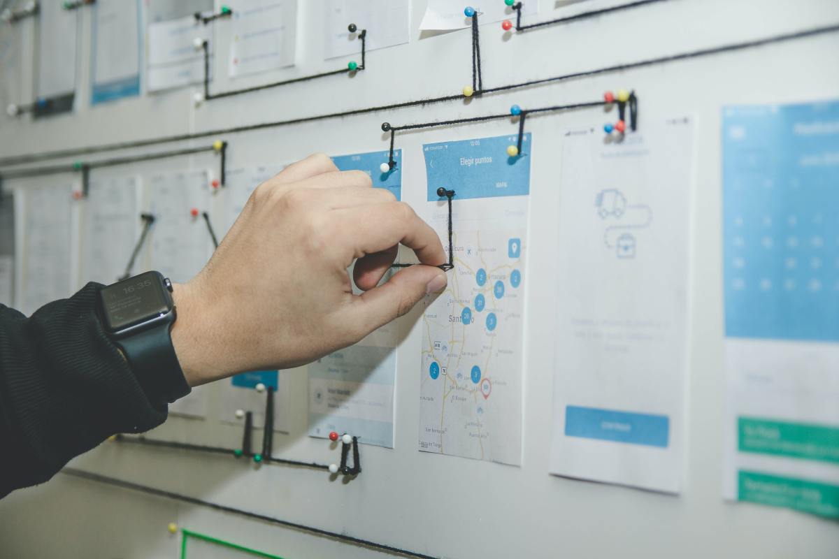 A person wearing a black smartwatch places a colorful pushpin into a white project board to connect printed user interface wireframes with black string.