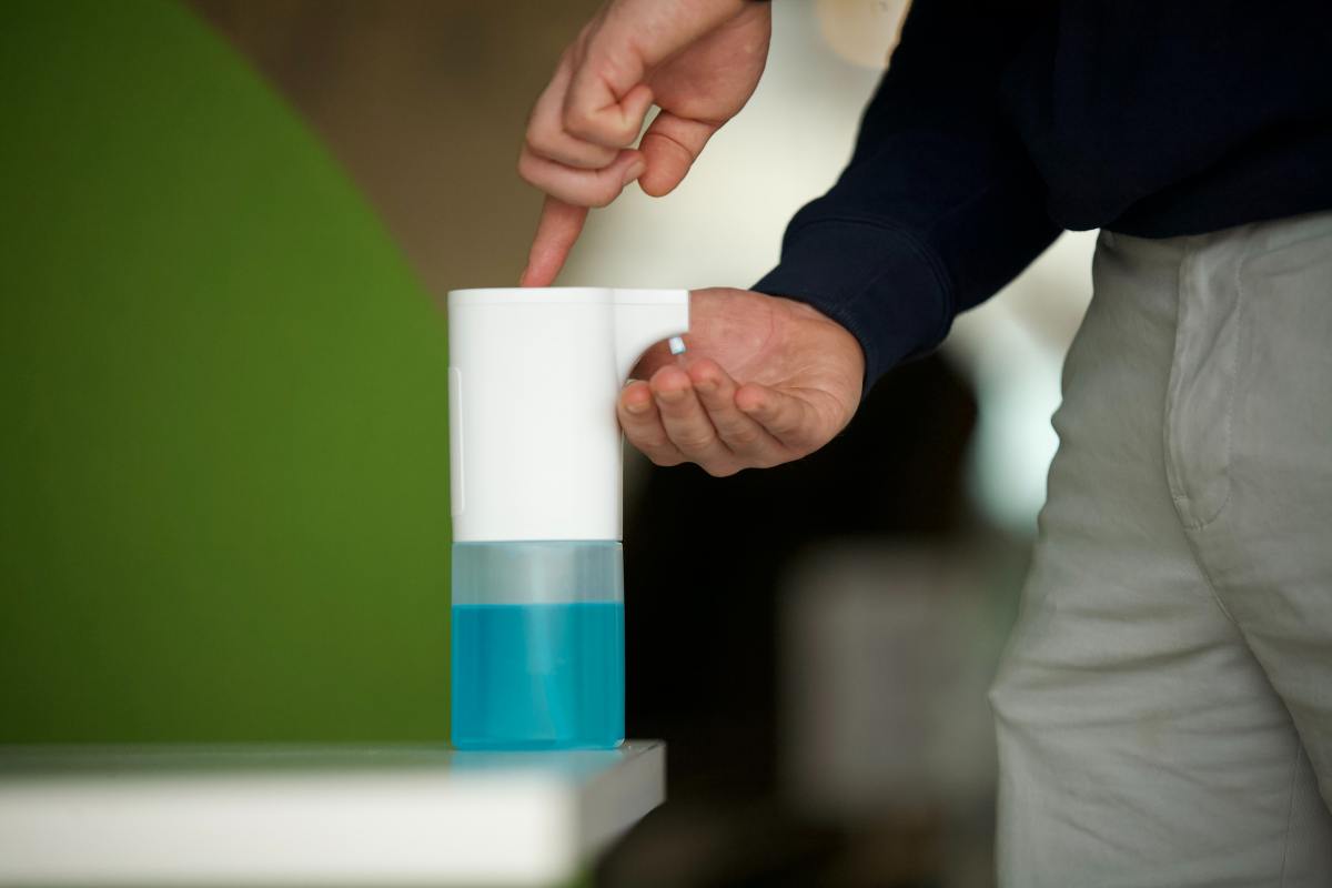 A person uses their index finger to press the top button of an automatic, white liquid dispenser with a clear base containing blue soap or hand sanitizer.