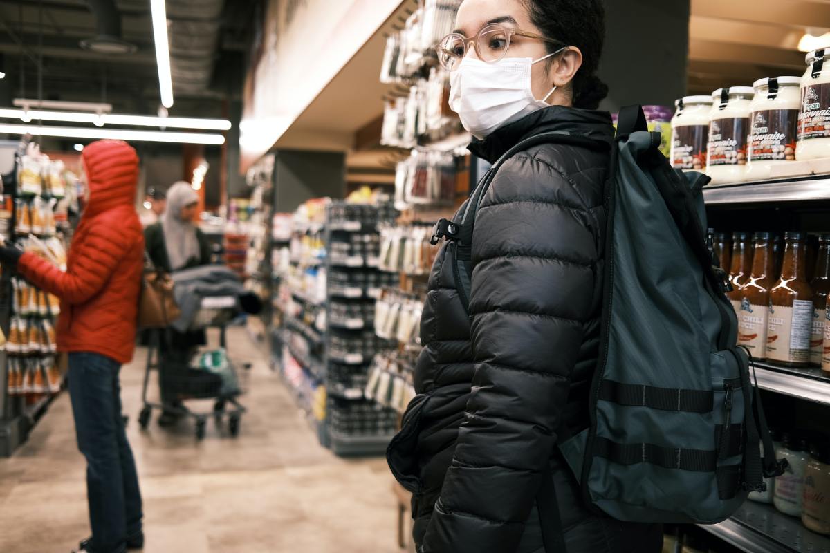A person wearing glasses, a white face mask, and a black puffer jacket looks sideways while standing in a grocery store aisle next to shelves of products.
