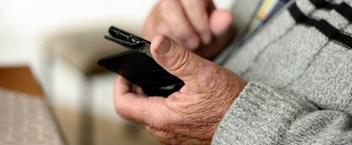 An elderly person's wrinkled hands hold and use a smartphone enclosed in a black wallet-style case.