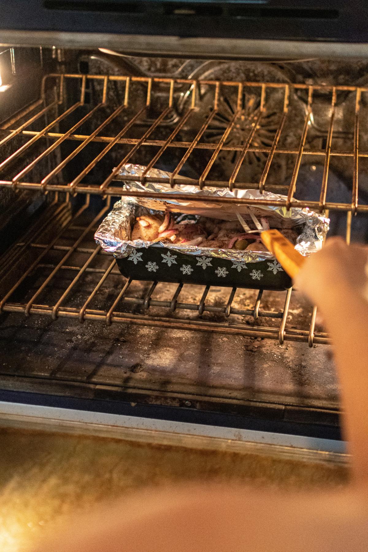 A hand reaches into a hot oven to peel back aluminum foil from a loaf pan using a fork, revealing food baking inside.
