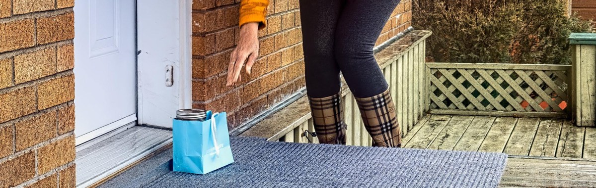 A person wearing plaid boots reaches down toward a small blue gift bag with a glass jar inside, placed on a doormat outside a brick house.