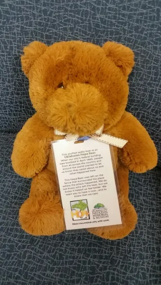 A brown, fluffy teddy bear sits upright against a textured blue background, wearing a white ribbon around its neck attached to an informational hanging tag from the Oklahoma City National Memorial & Museum.