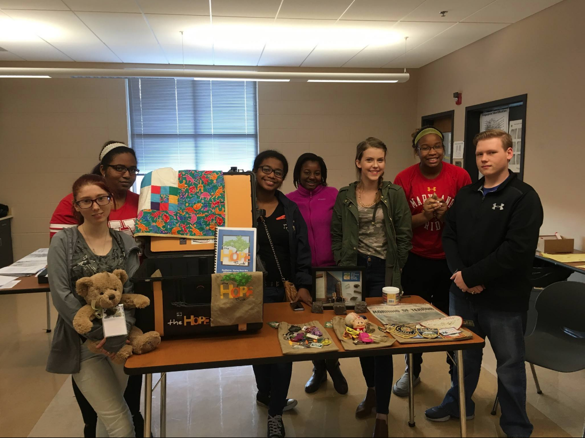 Group of students standing behind a display of personal items, keepsakes, and artwork as part of a community resilience and disaster recovery project in a classroom setting.